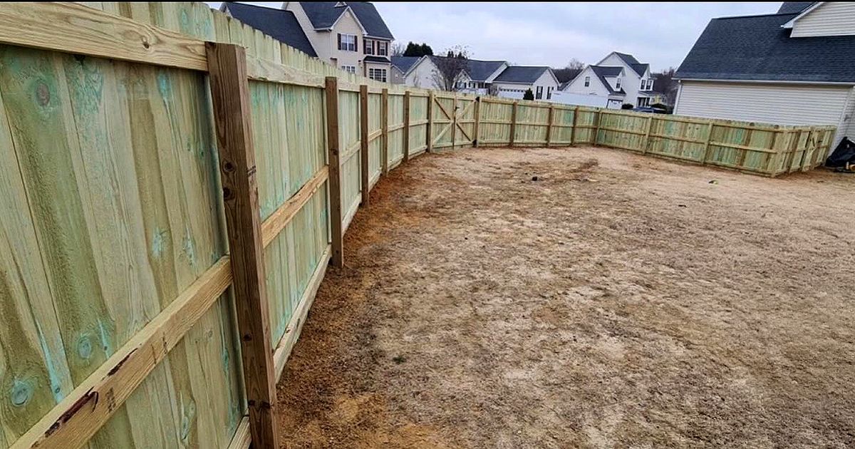 New wooden backyard fence enclosing a bare dirt yard with houses in the background