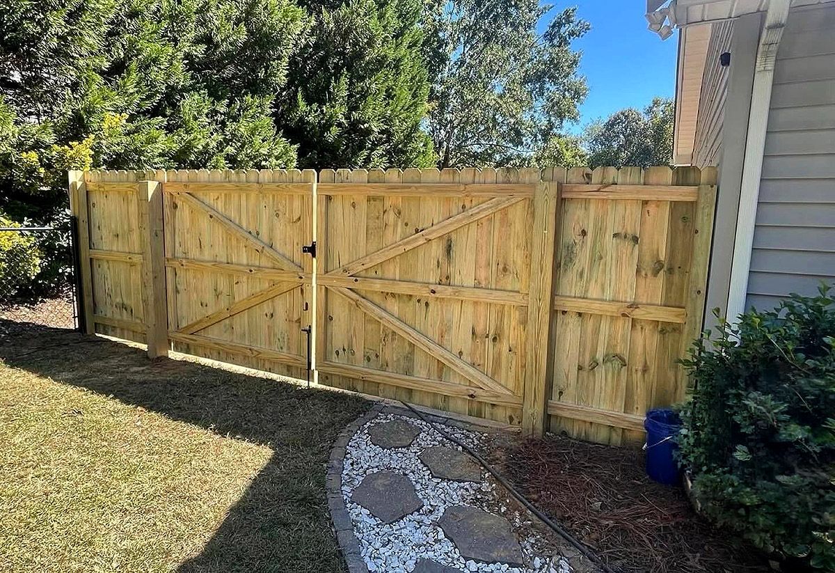 Wooden backyard fence gate beside a house, with trees and a stone path in sunlight