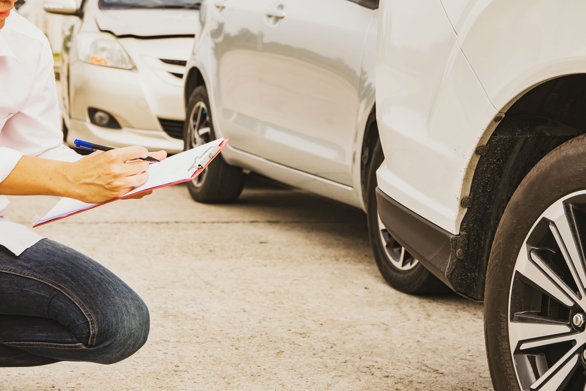 Person inspecting the side of a white car with a clipboard near a damaged wheel