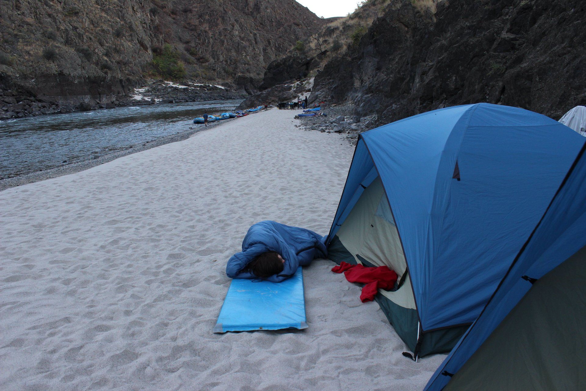 A person sleeping in a blue sleeping bag next to a tent on a beach on the Lower Salmon River