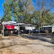 Covered parking area with several vehicles and a red car under metal canopies, trees in the background.
