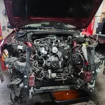 Car with hood open, front end dismantled, exposing engine and wiring in a garage