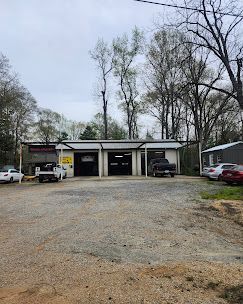 Small auto repair shop with three garage bays, parked vehicles, and a gravel lot under overcast trees.