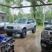 Silver SUV parked under a carport on a rainy driveway, with a black truck at right.