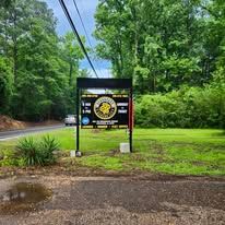 Black sign at a park entrance beside a wet road, surrounded by lush green trees and grass.