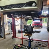 Car lifted on a garage hoist with a mechanic under it and tools on the floor, red SUV outside