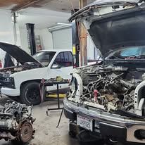Auto repair shop with several vehicles and engines disassembled, including a black truck and white pickup.