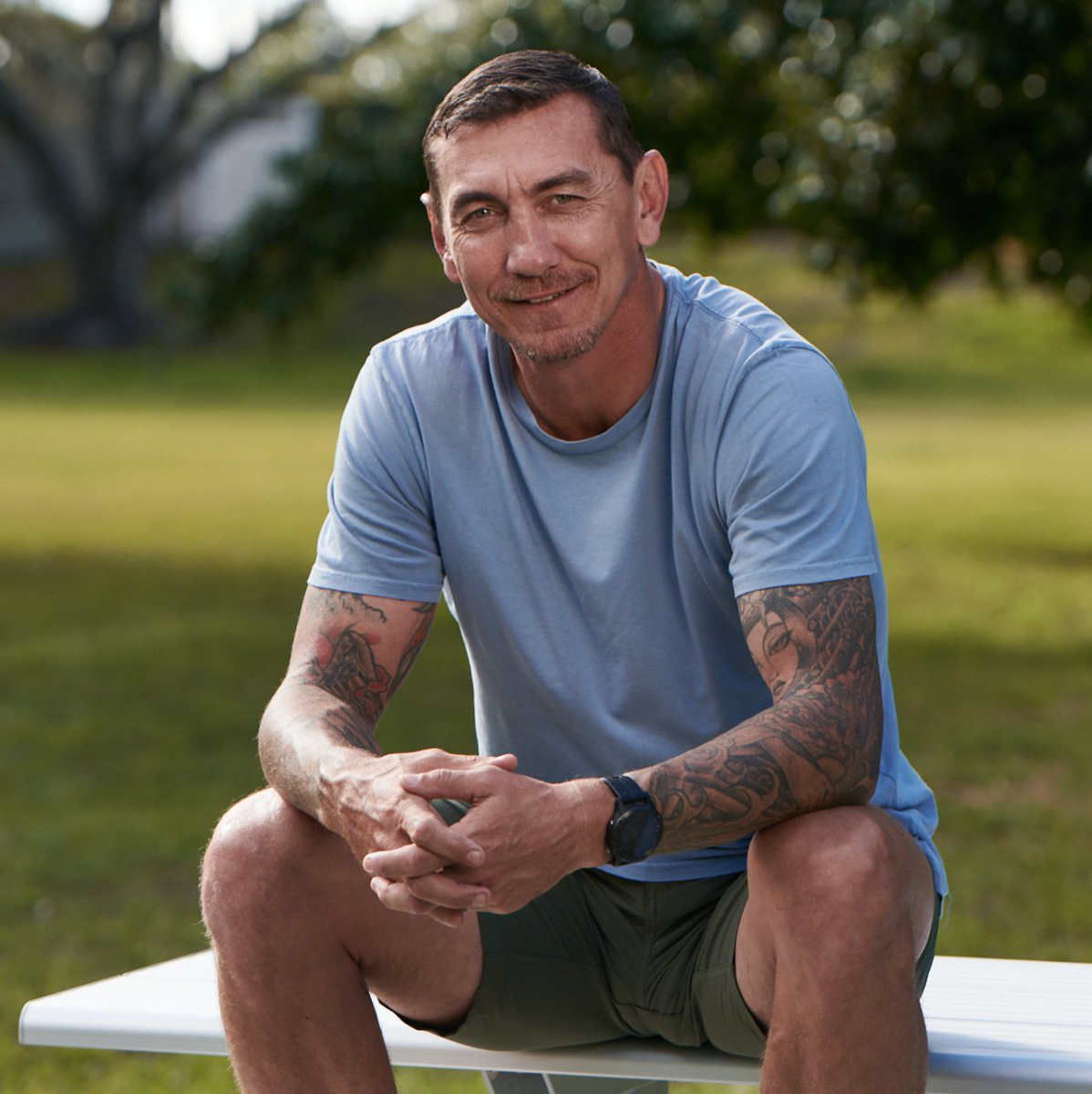 A man with tattoos is sitting on a bench in a park
