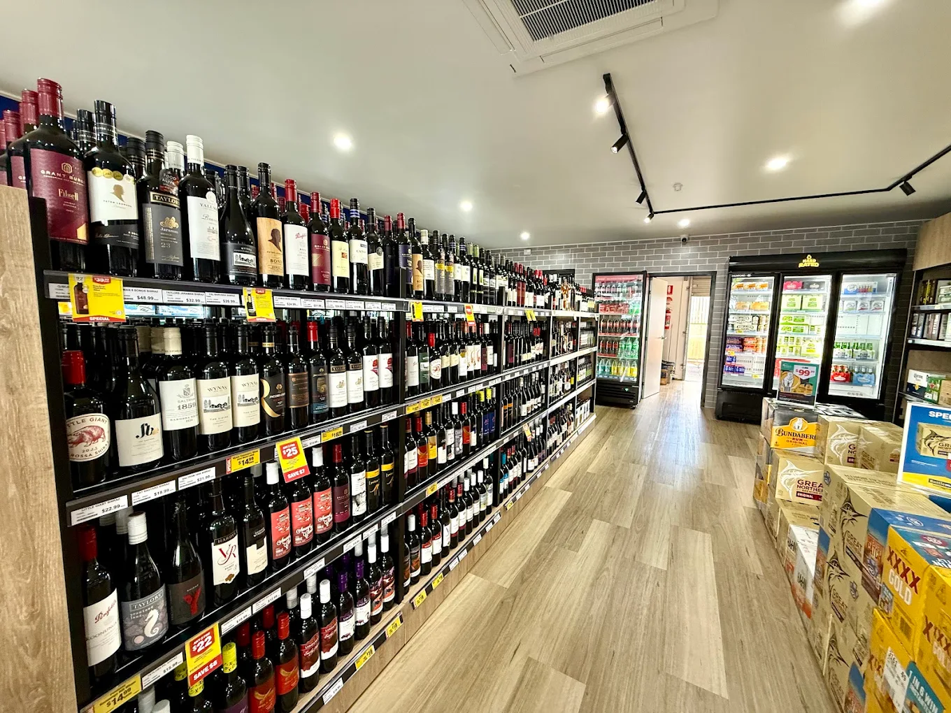 A convenience store aisle with wine bottles on shelves and refrigerated drinks at the back.