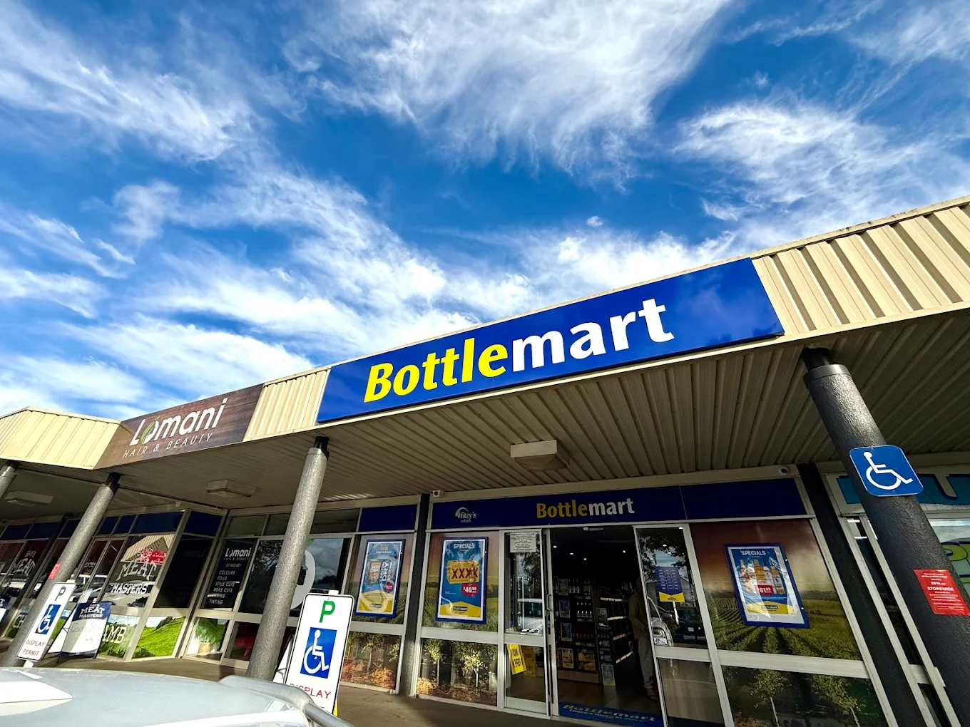 Bottlemart storefront with blue sign under a bright sky, with window posters and accessible parking sign
