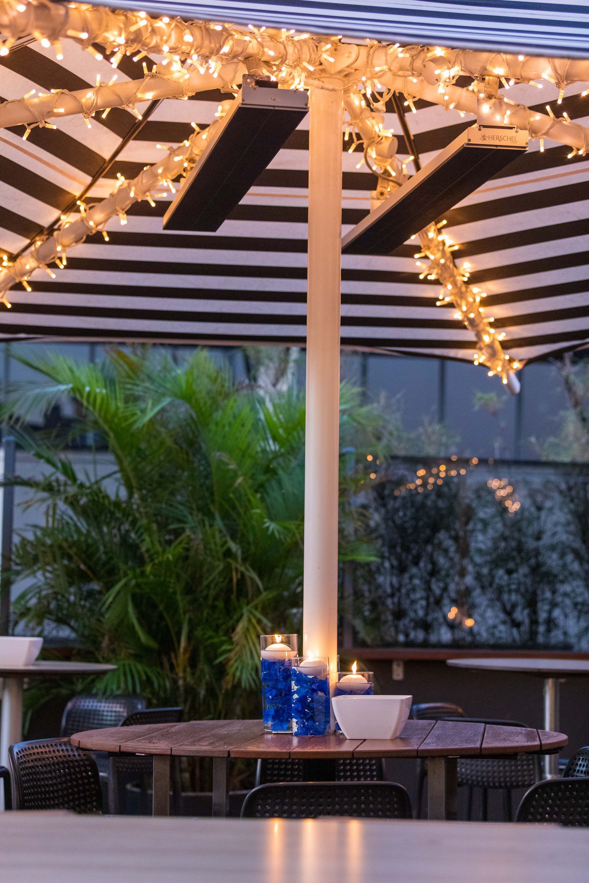 String lights over an outdoor patio table with striped canopy and potted plants at dusk