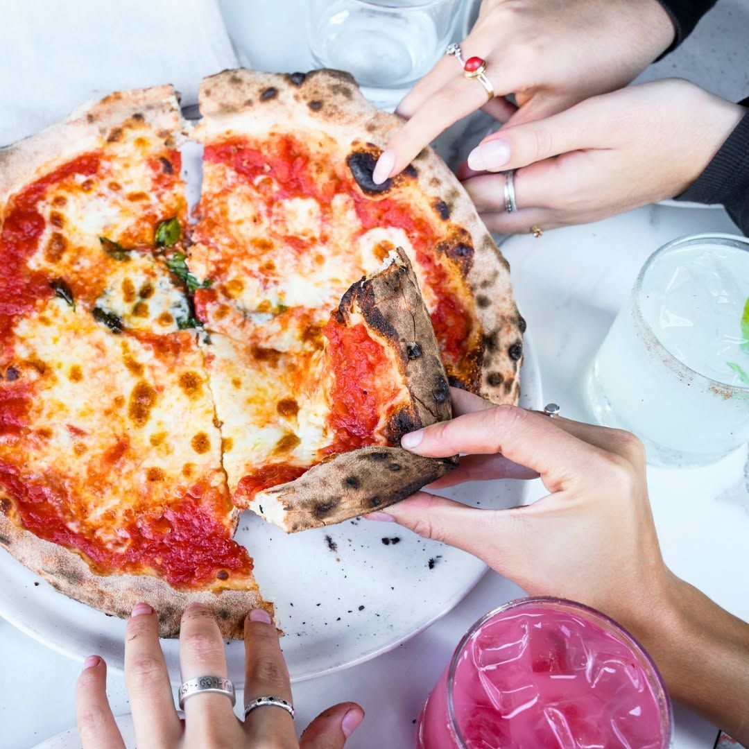 Hands reaching for slices of cheese pizza on a table with drinks