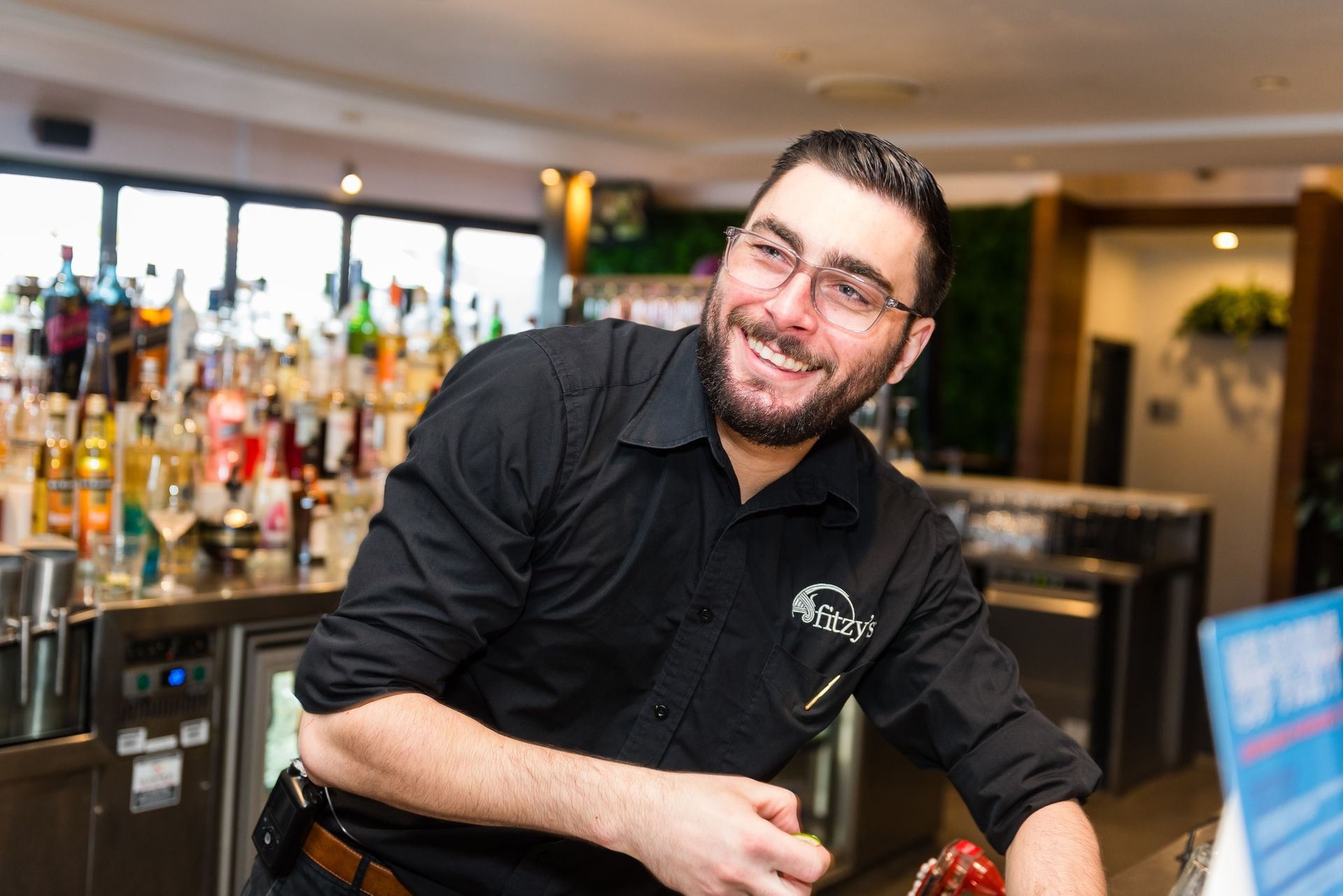Smiling bartender in a black shirt leaning on a bar with bottles and glasses behind him