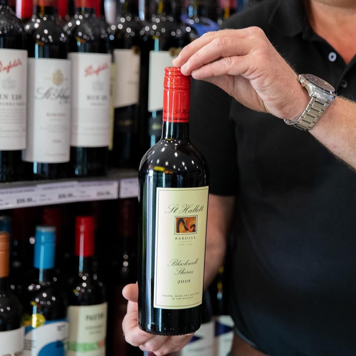 A person holds up a red wine bottle in a store, surrounded by wine bottles on shelves.