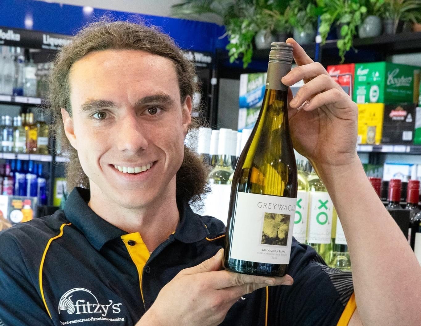 Man in Fitzys uniform holding a bottle of white wine, smiling in a store.