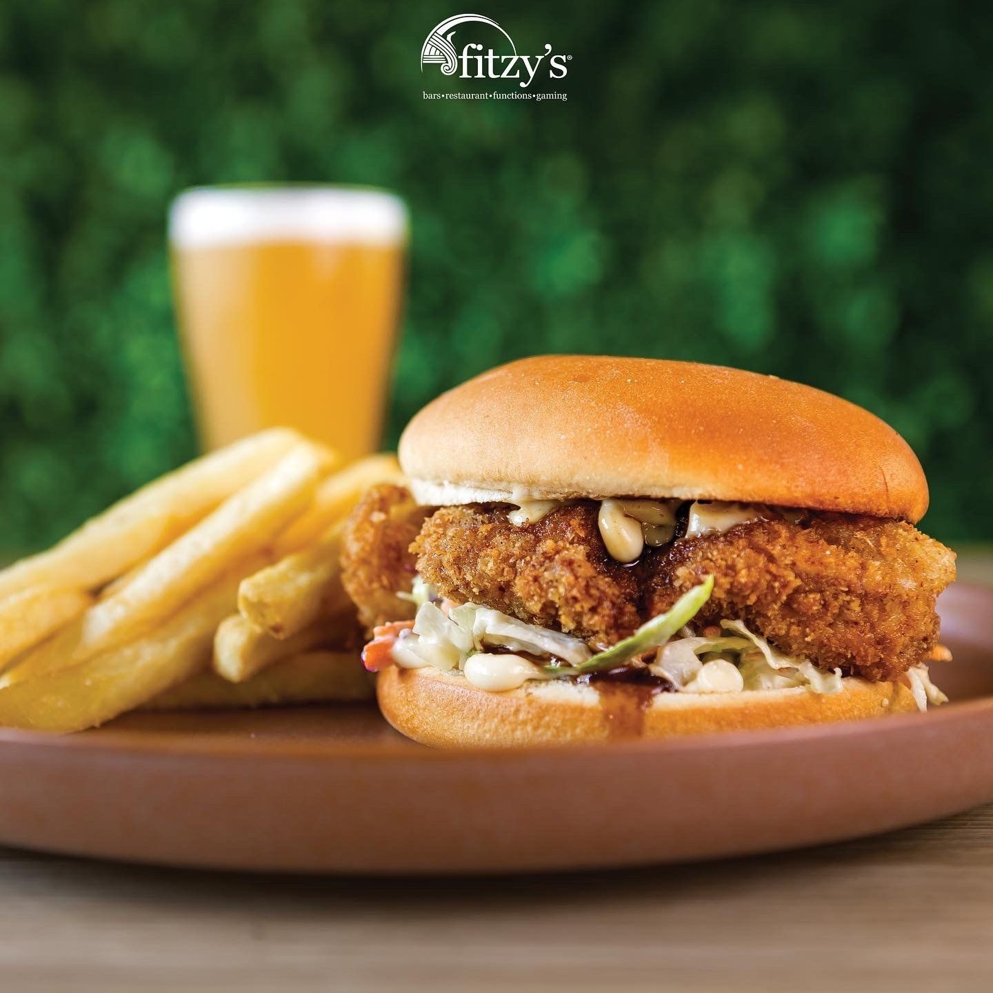 A fried chicken sandwich and french fries on a plate with a glass of beer in the background