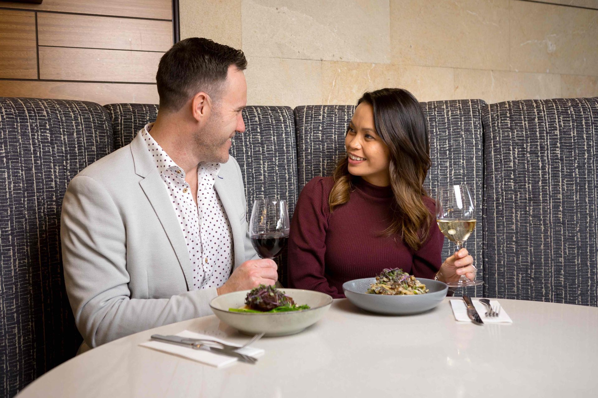 A man and a woman are sitting at a table eating food and drinking wine.