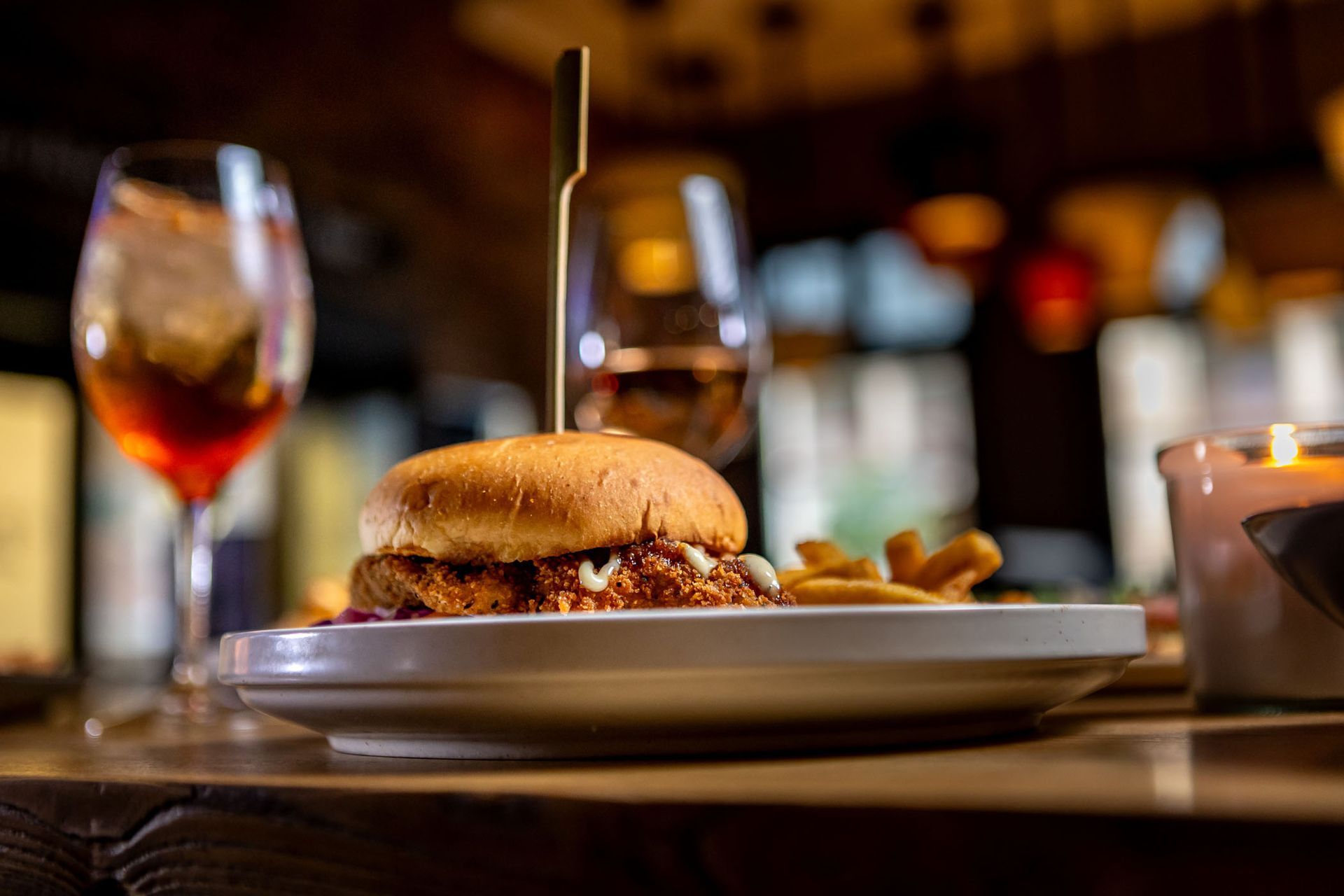 Burger on a plate with fries, a drink, and a candle on a wooden table in a restaurant.