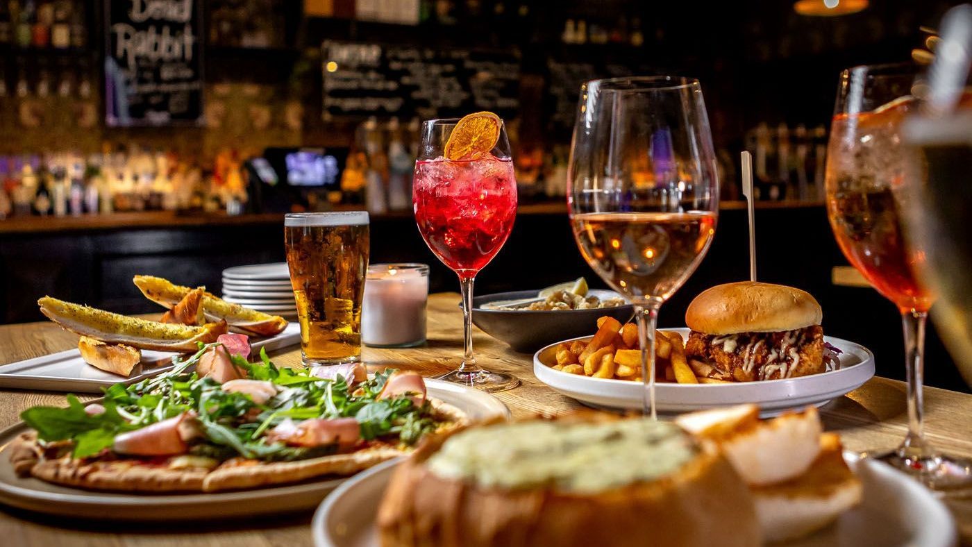Table with drinks (cocktails, wine, beer) and food in a bar: pizza, fries, burger, soup, and appetizers.