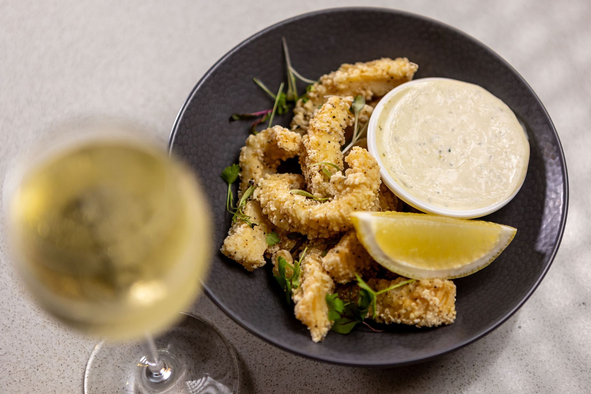 Fried calamari with dipping sauce and lemon wedge on a black plate, with a glass of white wine.
