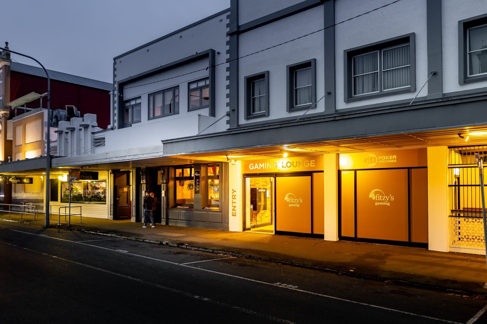 Lit storefronts in a row of white buildings along a street at dusk