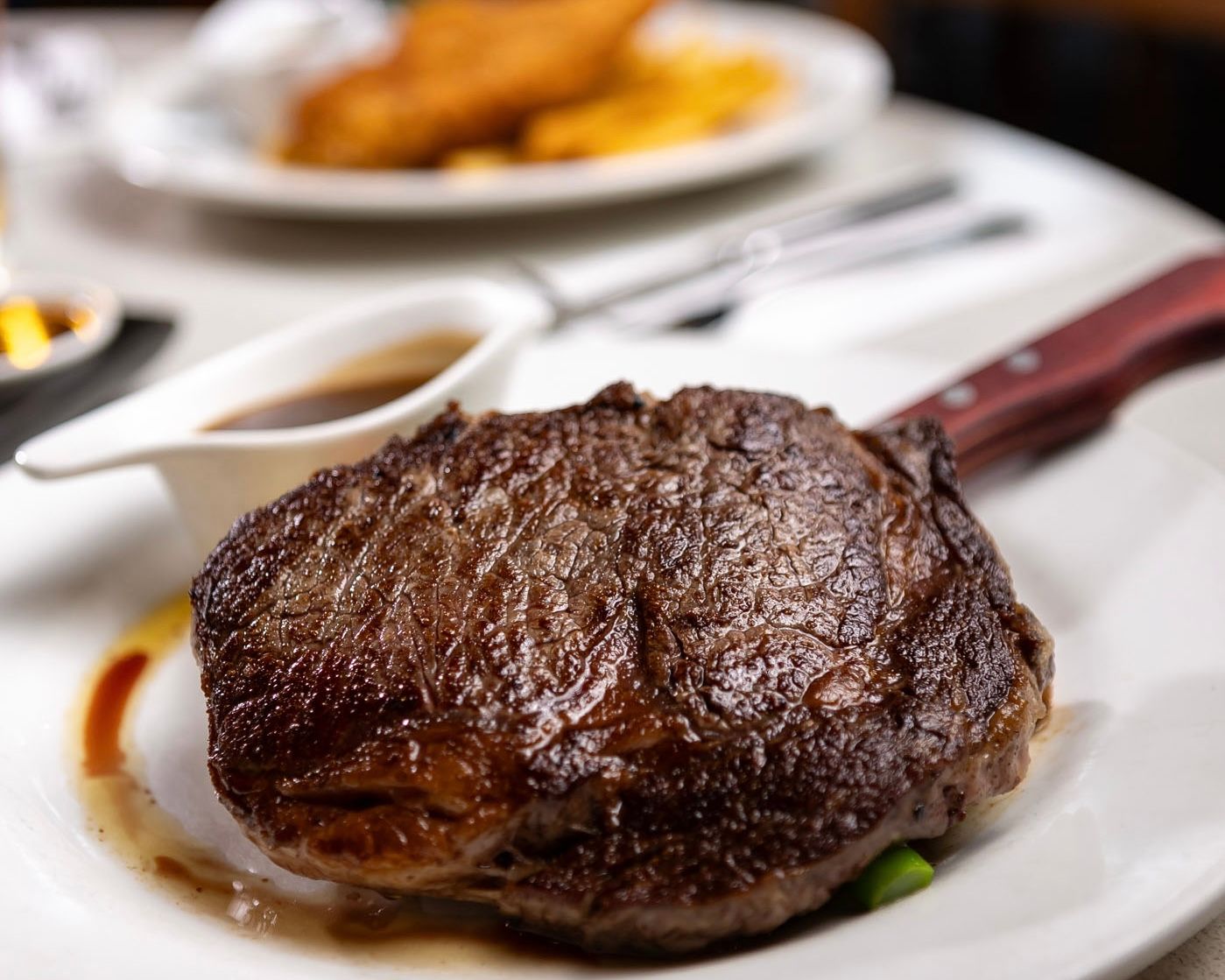 Grilled steak on white plate with gravy boat, knife, and blurred plate of fries.