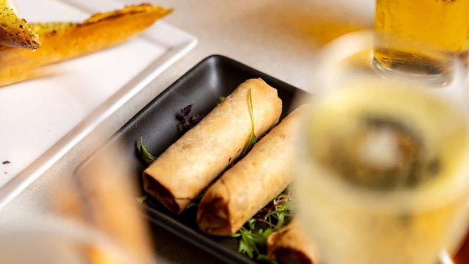 Two spring rolls on a black plate, with a drink in the foreground and garlic bread in the background.
