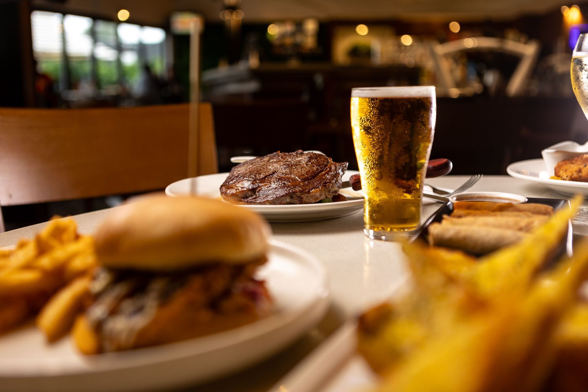 Table with a burger, steak, fries, and a glass of beer. Restaurant setting, blurred background.