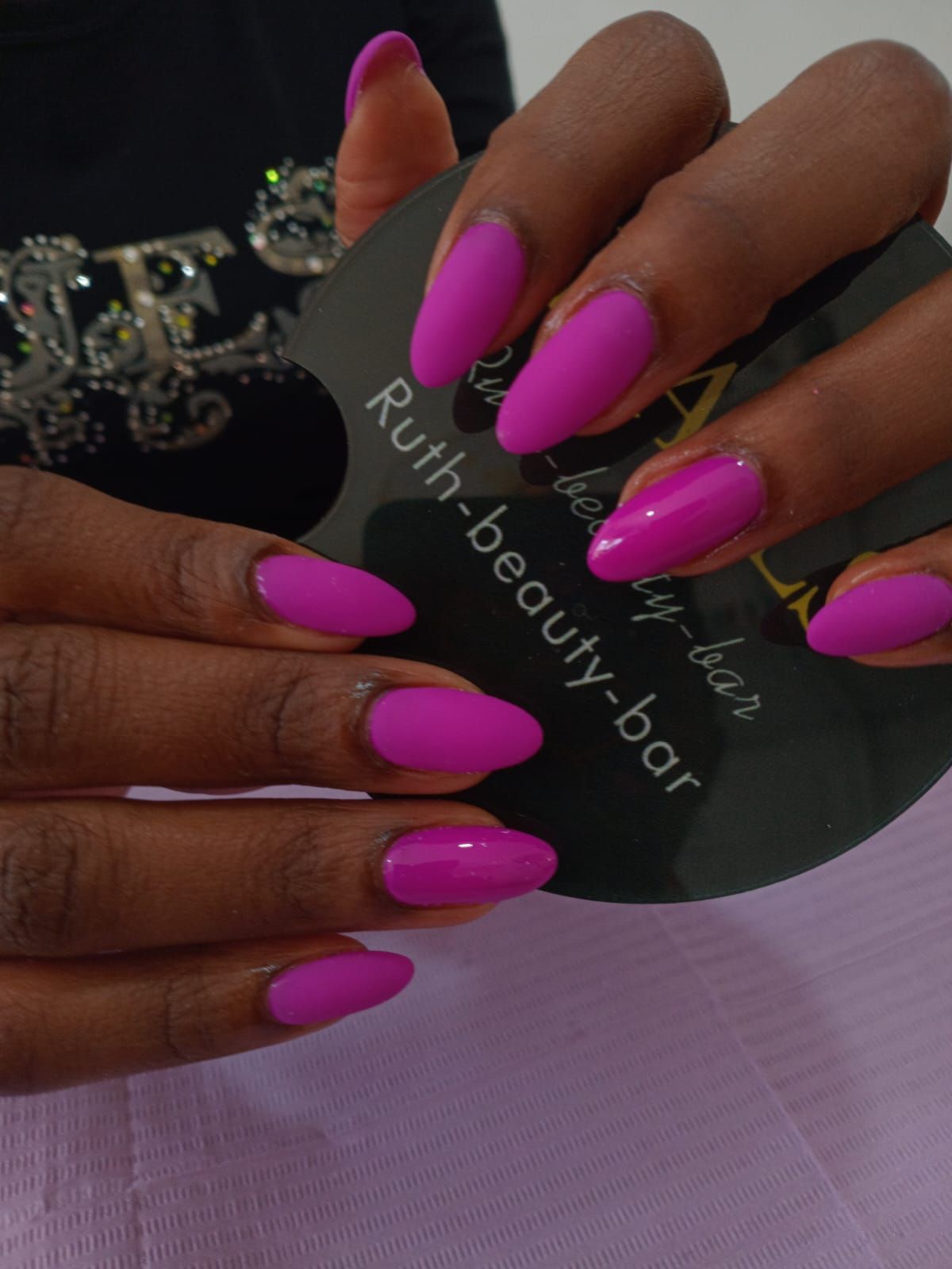 Two hands display almond-shaped nails painted in a vibrant magenta, holding a black 