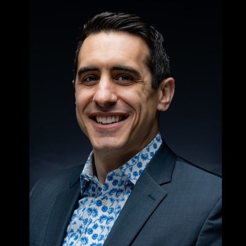 Smiling man in a navy suit and patterned blue shirt against a dark background.