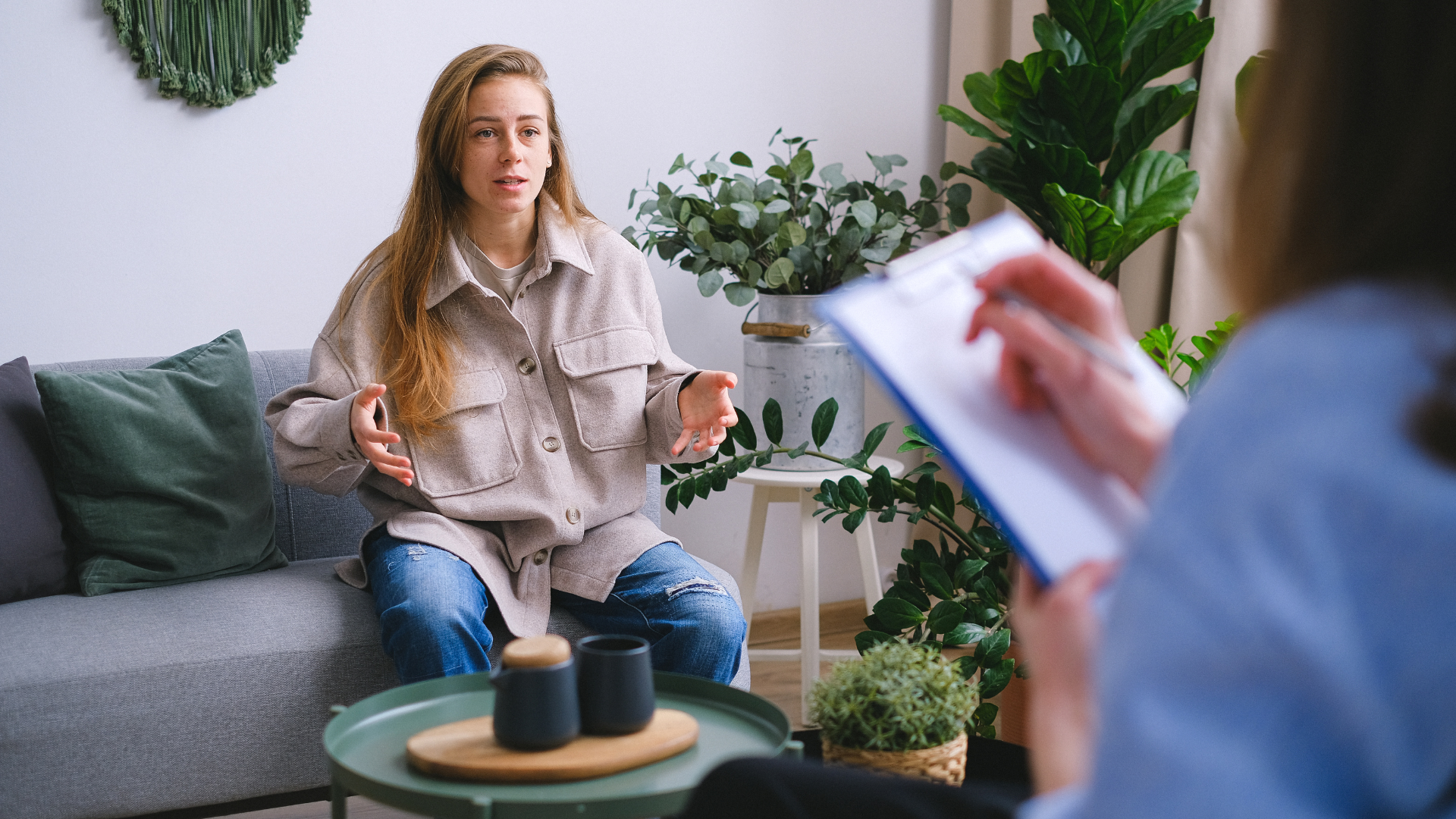 Woman on a couch gesturing, talking to a person holding a clipboard, indoors.