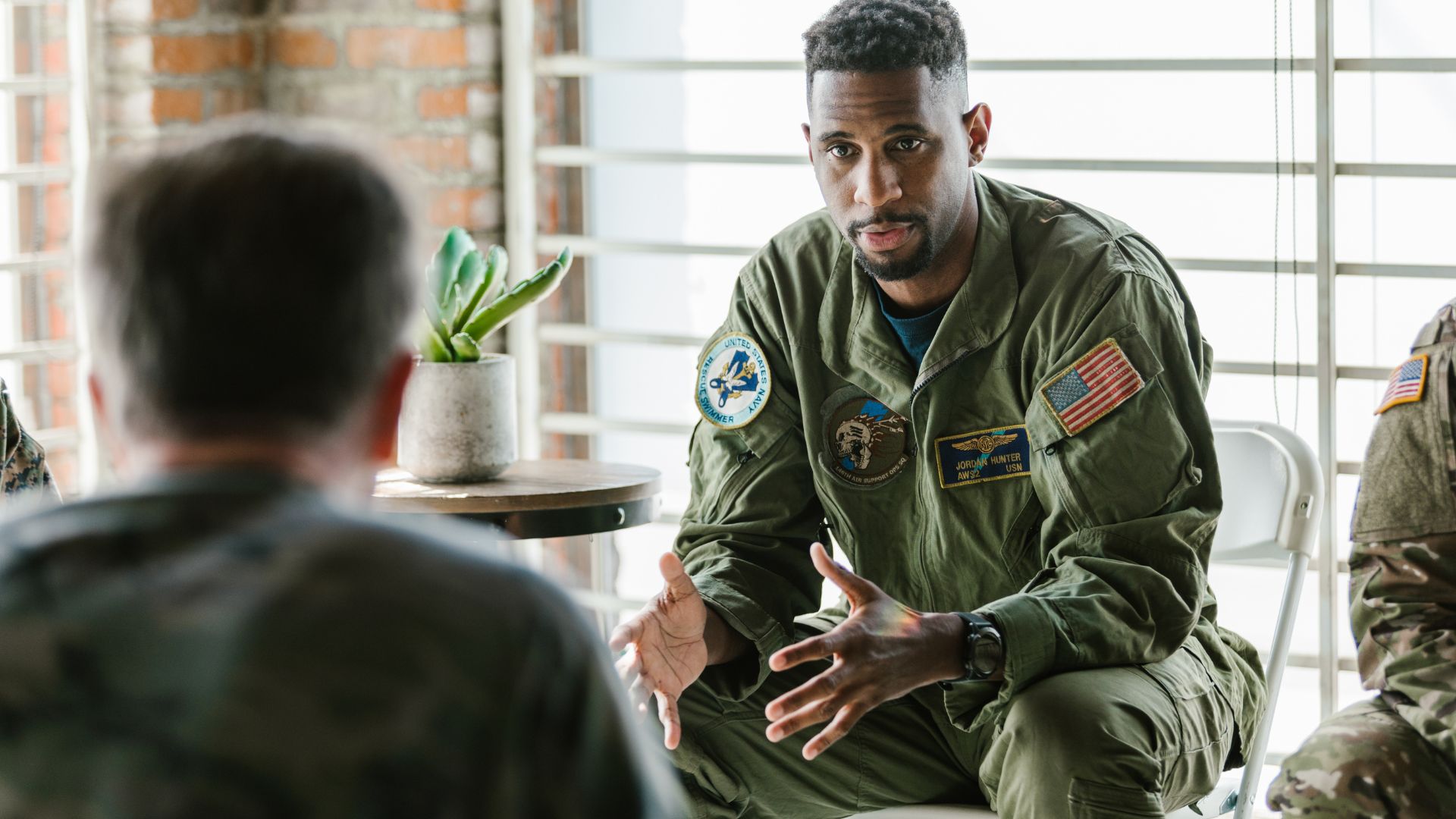 Military personnel in a support group, one speaking with hands, indoors, around small table.