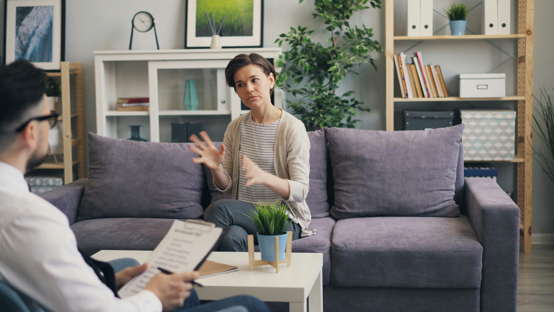 Woman gesturing, talking to person holding a clipboard, seated on sofa. Therapy session in a living room.