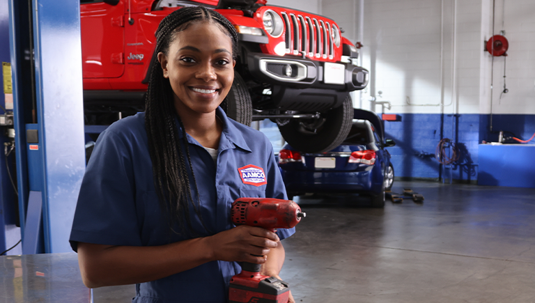 AAMCO mechanic holds a tool, smiling in a garage with a red jeep raised on a lift.
