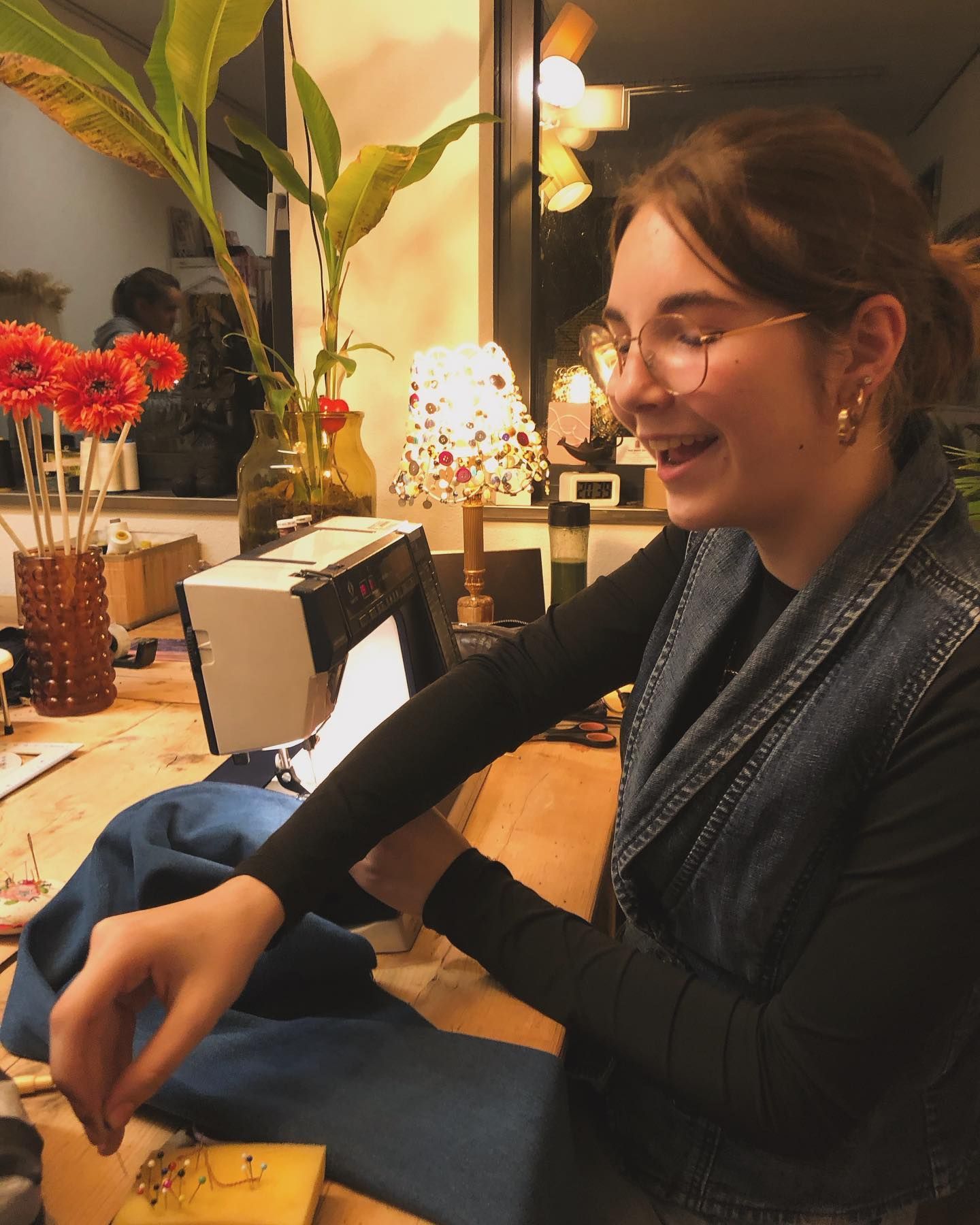 Een vrouw met een bril glimlacht terwijl ze aan een tafel met een naaimachine en bloemen naait, verlicht door een lamp.