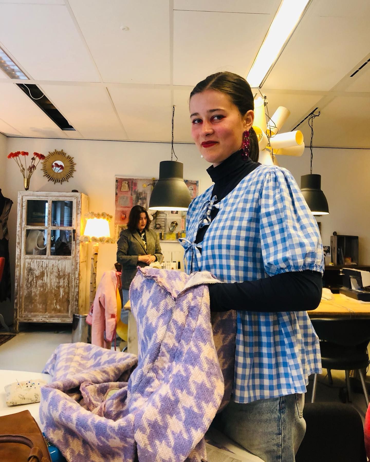 Een vrouw met een paarse jas met patroon staat in een kamer met een andere persoon, kasten en lampen.