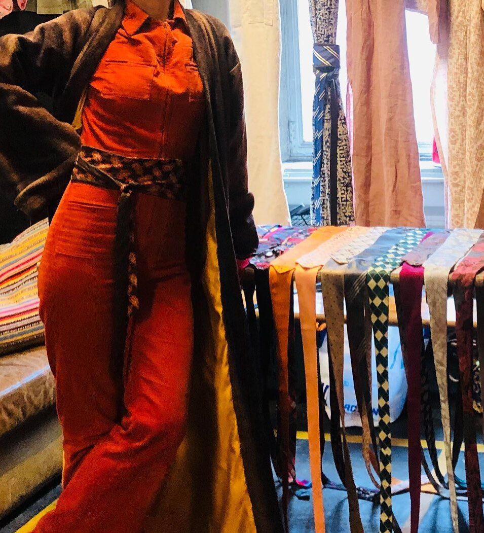 Een vrouw in een oranje jumpsuit, een donkere trui en een riem met patroon staat bij een tafel met riemen.