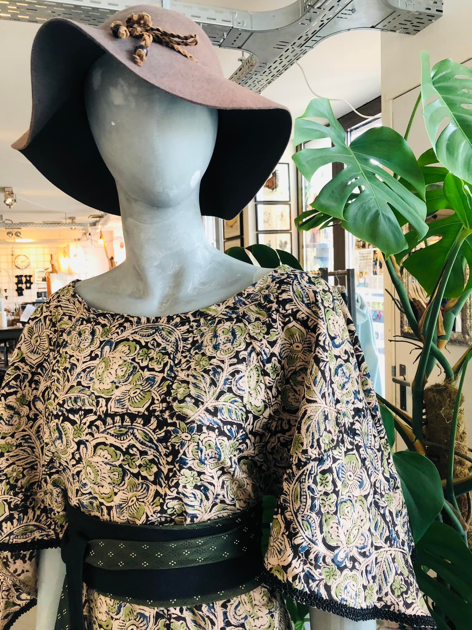 Een mannequin in een jurk met print, een hoed met brede rand en een zwarte riem. De mannequin staat tentoongesteld in een winkel met planten en kunstwerken.