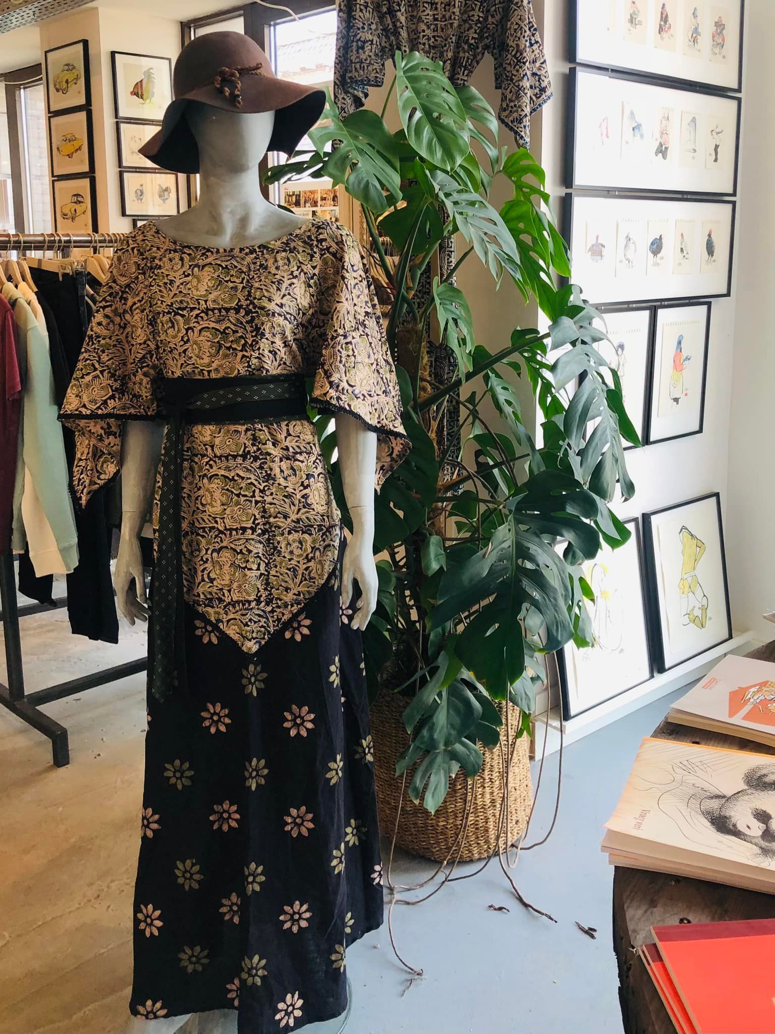 Een mannequin in een vintage jurk en hoed, tentoongesteld in een winkel met kunst en een potplant.