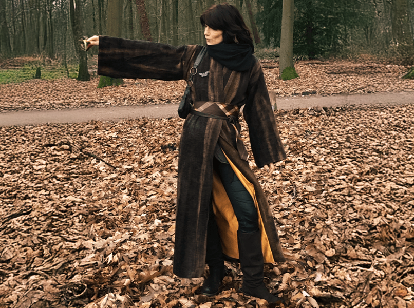 Vrouw in een bruine jas, wijzend, op een bospad bedekt met gevallen bladeren.