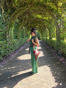 Een vrouw in een groene outfit staat onder een met bladeren begroeide boog en houdt bloemen vast. Een stenen pad leidt in de verte.