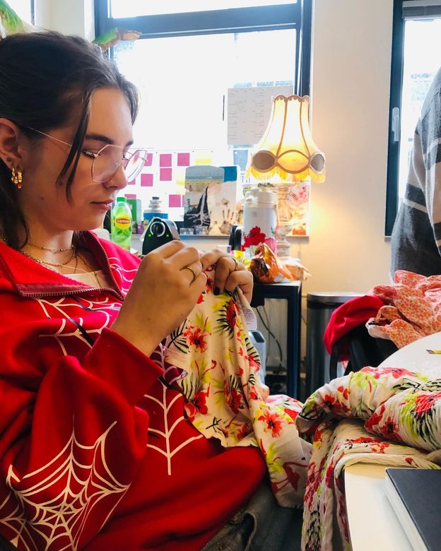 Vrouw in rode sweater naait bloemenstof. Lichte kamer met raam, lamp en naaibenodigdheden.