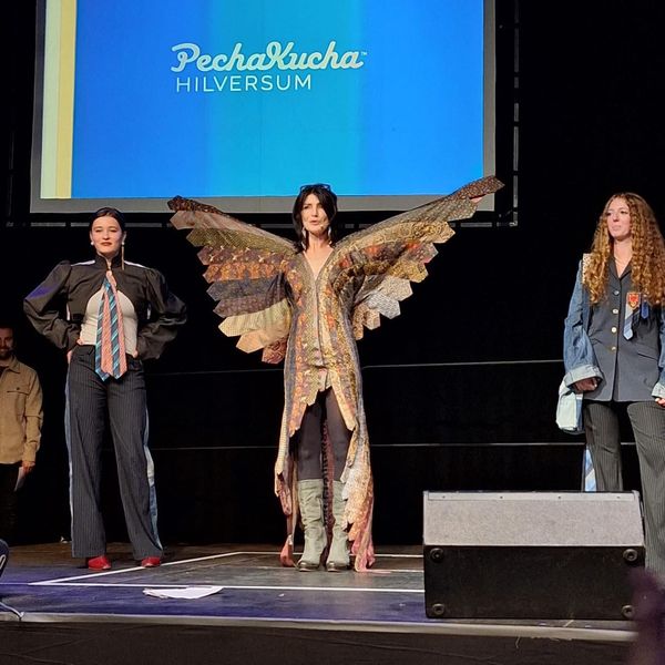 Drie mensen staan ​​op een podium voor een scherm met een logo. Eén van hen draagt ​​vleugels, de anderen pakken.