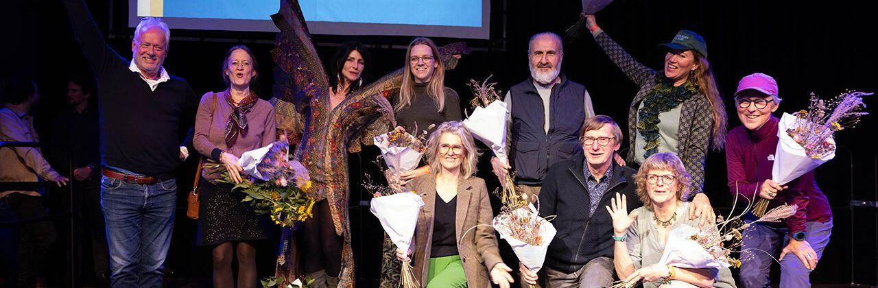 Een groep mensen staat op een podium met bloemen in hun handen. Sommigen glimlachen, anderen kijken naar het publiek. Donkere achtergrond.