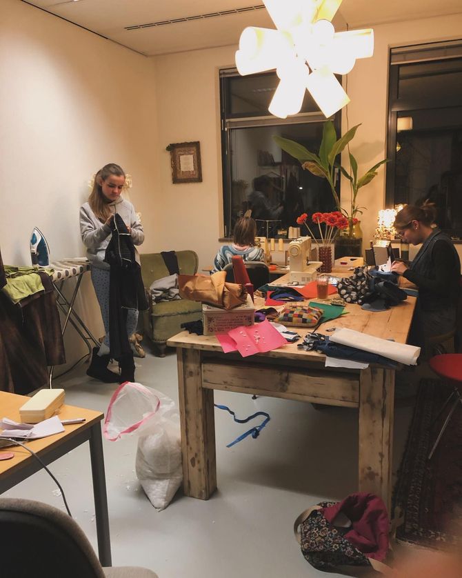 Mensen naaien in een goed verlichte werkplaats met knutselspullen. Eén persoon staat, anderen zitten aan tafels.