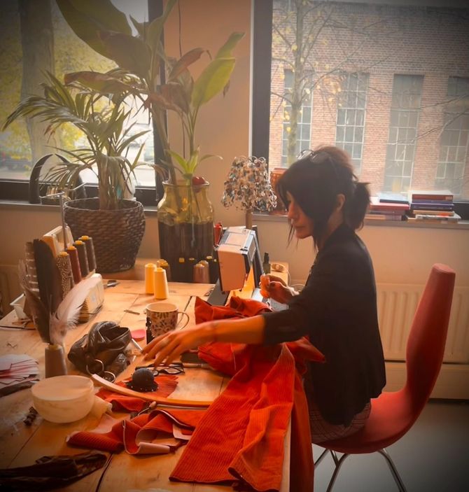 Een vrouw naait aan een bureau bij een raam met planten, verlicht door natuurlijk licht. Rode stof en naaibenodigdheden liggen op tafel.