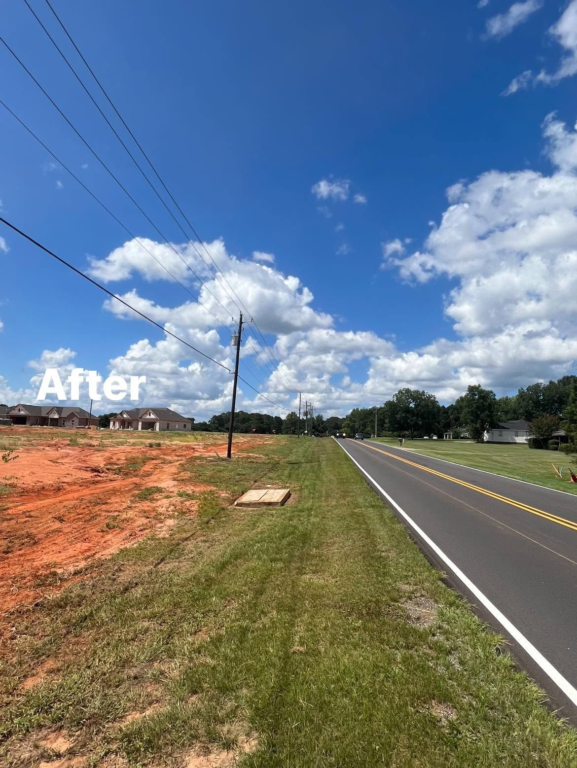 Rural roadside beside cleared red dirt lot under bright blue sky with clouds