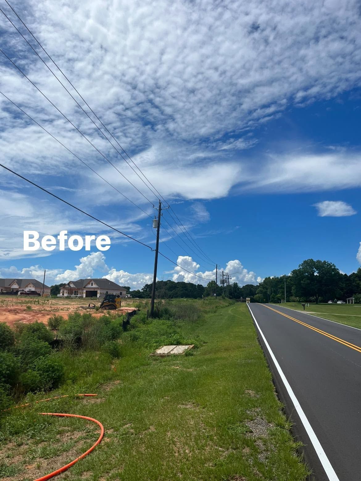 Rural roadside with grass verge, cloudy blue sky, and a marked road beside new construction site.