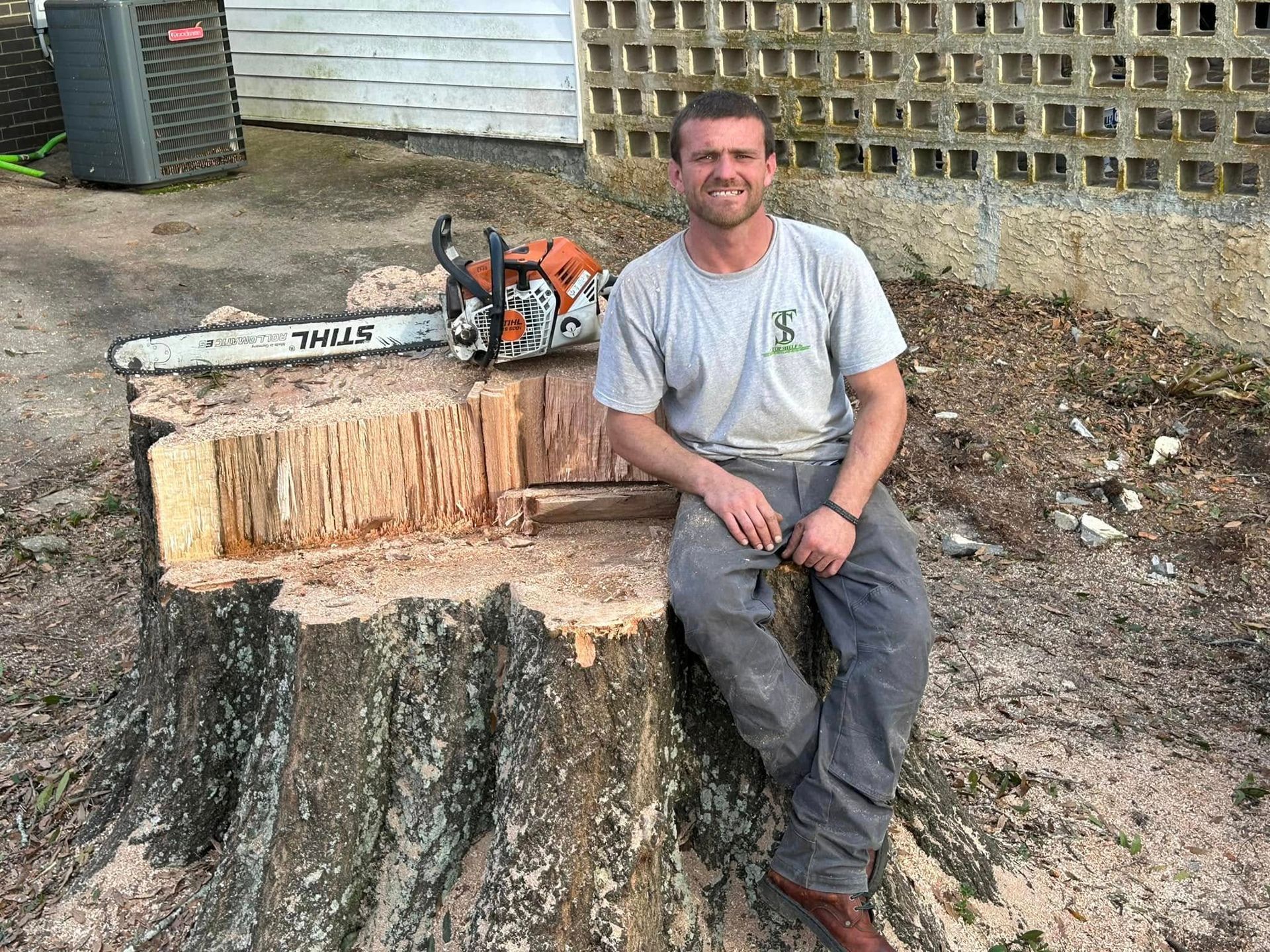 Man sitting on a large tree stump beside a chainsaw in a yard, smiling at the camera.