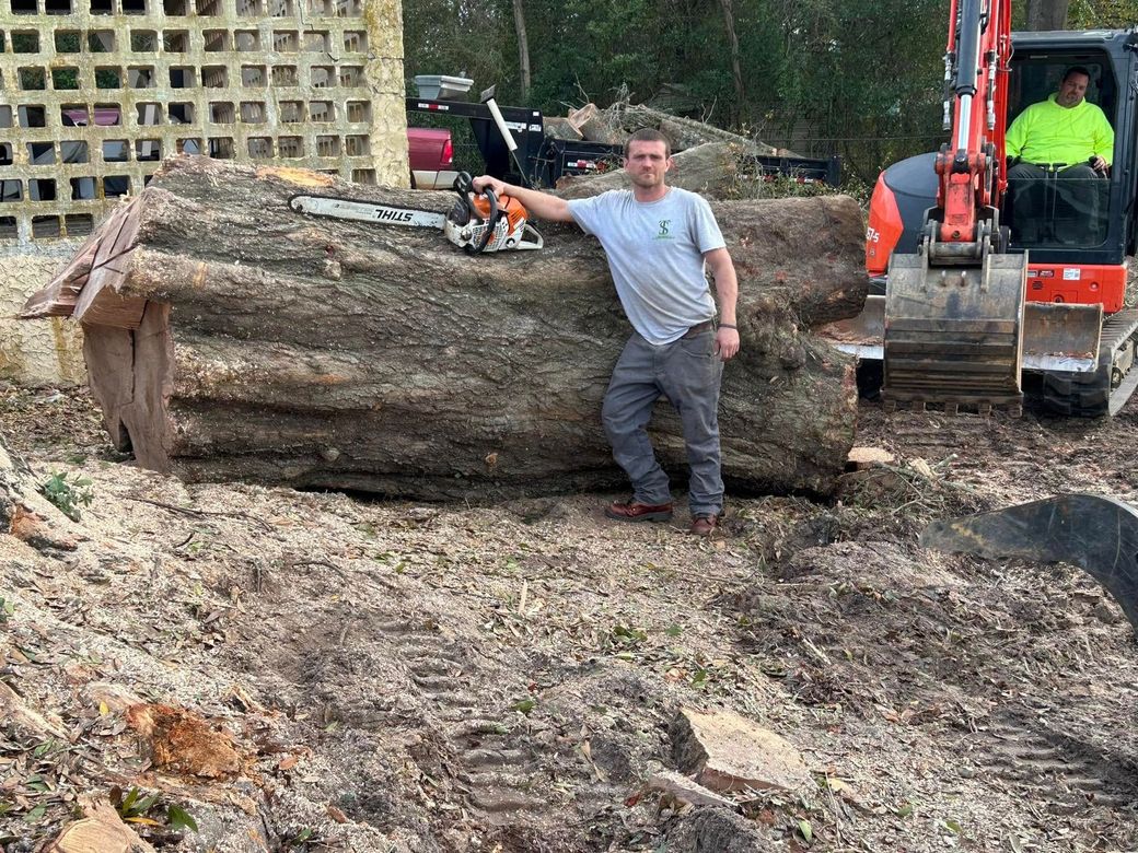 Man posing beside a huge fallen log at a worksite, with a small excavator nearby.